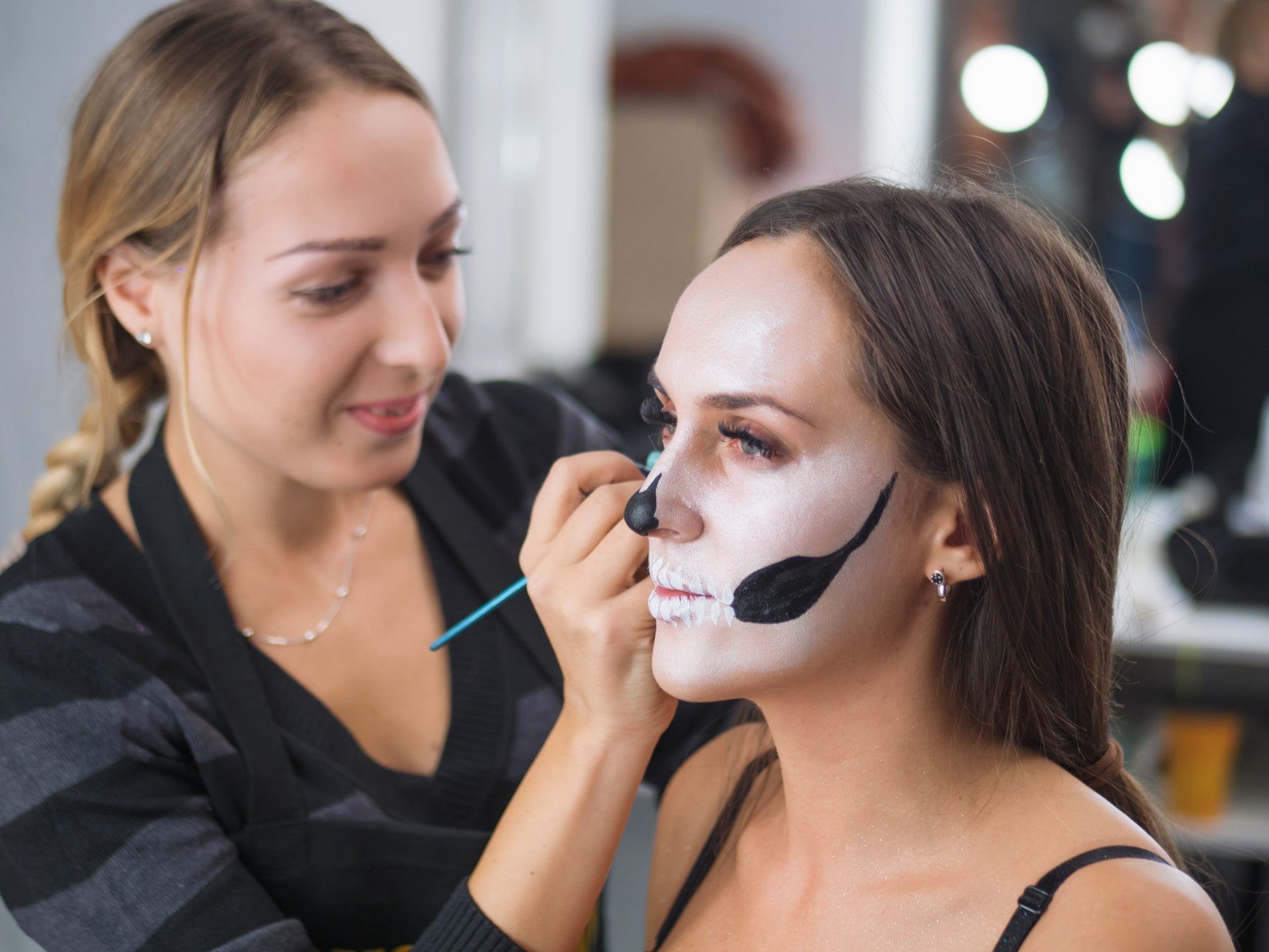 A woman putting white and black Halloween makeup on another woman